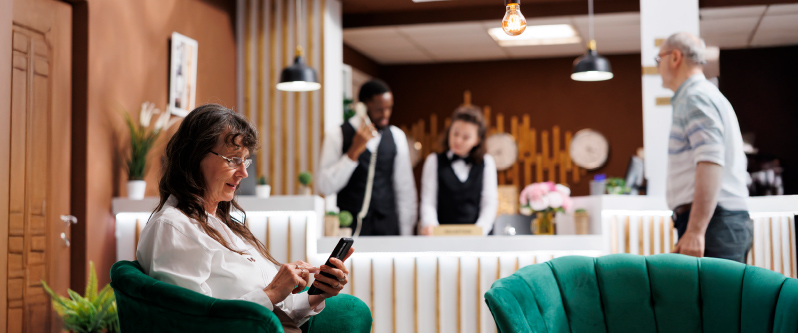 Woman using her smartphone in a hotel lobby while guests interact with reception staff in the background